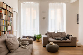 Modern living room with neutral sofas, bookshelf, pouf, and large windows with sheer curtains.